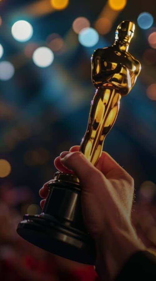 Close-up of a hand holding a gold Academy Award-style trophy against a blurred background of stage lights and a cheering audience.