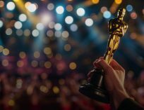 Close-up of a hand holding a gold Academy Award-style trophy against a blurred background of stage lights and a cheering audience.