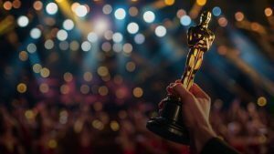 Close-up of a hand holding a gold Academy Award-style trophy against a blurred background of stage lights and a cheering audience.
