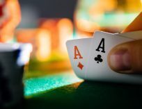 Close-up of a poker hand holding ace of diamonds and ace of clubs over a green felt table, with blurred casino chips in the background.