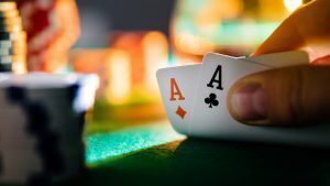 Close-up of a poker hand holding ace of diamonds and ace of clubs over a green felt table, with blurred casino chips in the background.