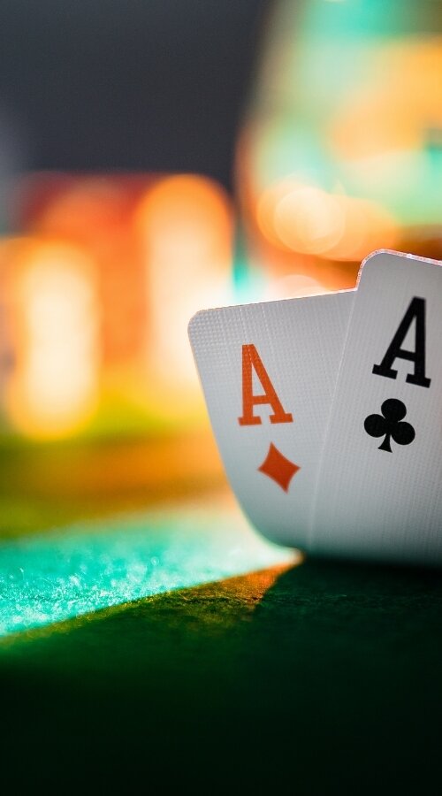 Close-up of a poker hand holding ace of diamonds and ace of clubs over a green felt table, with blurred casino chips in the background.