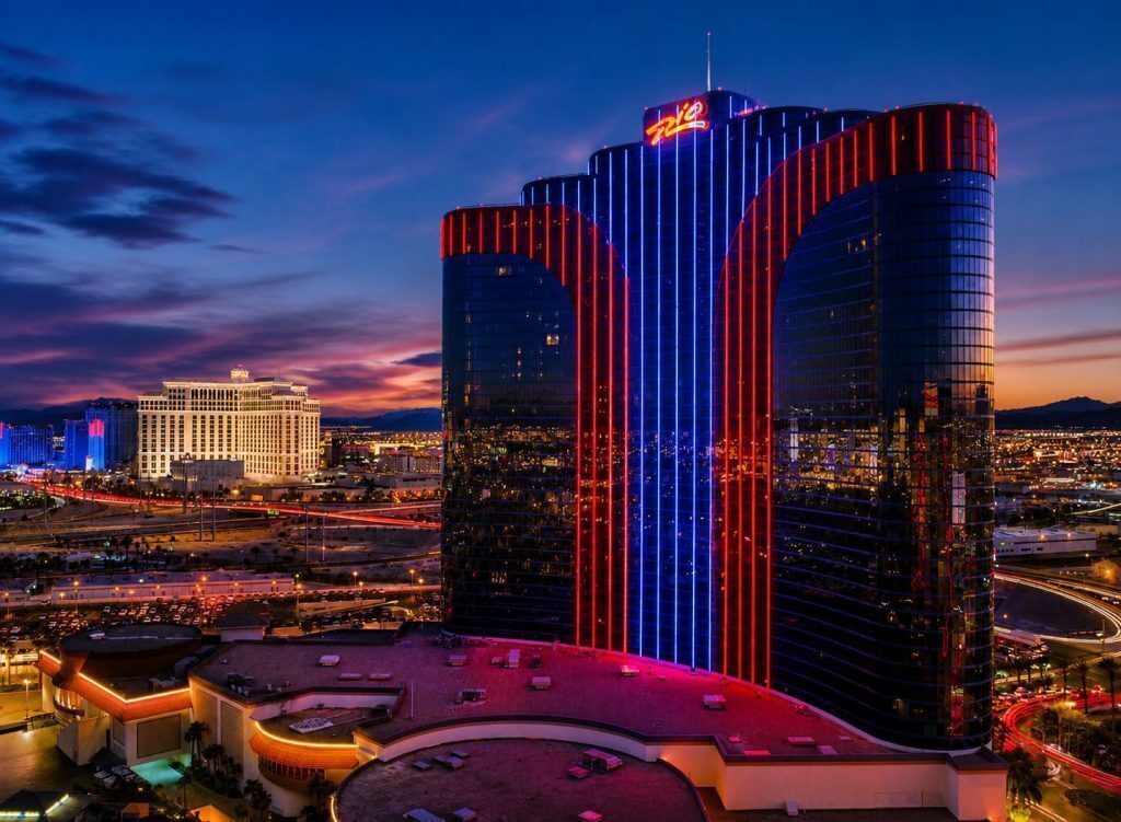 Rio hotel and casino in Las Vegas at dusk with red and blue neon lights, city skyline, and surrounding roads illuminated at night