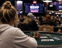 Female poker player in a hoodie seated at a tournament table with chips and cards in focus, surrounded by a busy WSOP-style poker room in the background