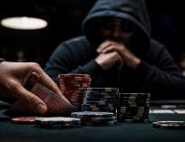 Close-up of a poker hand discreetly holding cards behind stacked chips at a dimly lit table, with a shadowy figure watching in the background, suggesting suspicious or unethical play.