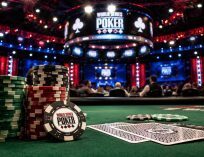 Wide cinematic view of a World Series of Poker tournament floor with poker chips, cards, and a green felt table in the foreground under bright arena lighting at a televised poker event.