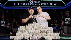 Two poker tournament winners pose with a championship belt behind a large stack of cash and poker chips at the final table.